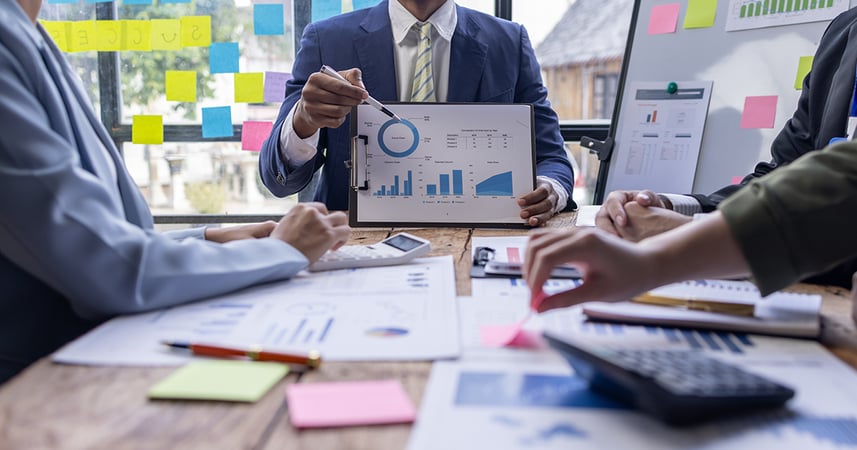 a man showing graphs and charts in a busy conference room table full of people and papers
