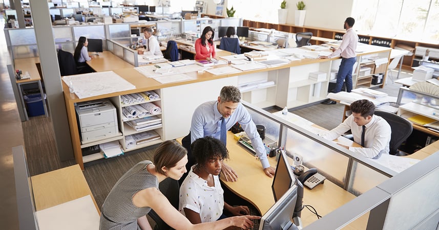 busy office with people working at open desks with computers and multiple screens