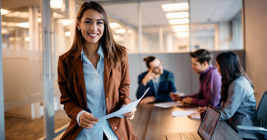 Woman in conference room holding a printed piece of paper. Her colleagues are having a discussion behind her at the table. 