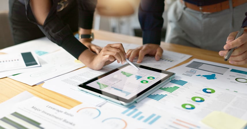 Employees reviewing budgeting graphs and charts on printed papers and an electronic tablet located on a conference room table.