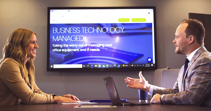 two businesspeople sitting at a conference room table with an interactive display in the room showing a presentation