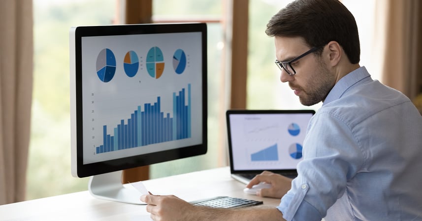 man looking at three computer screens filled with graphs and charts
