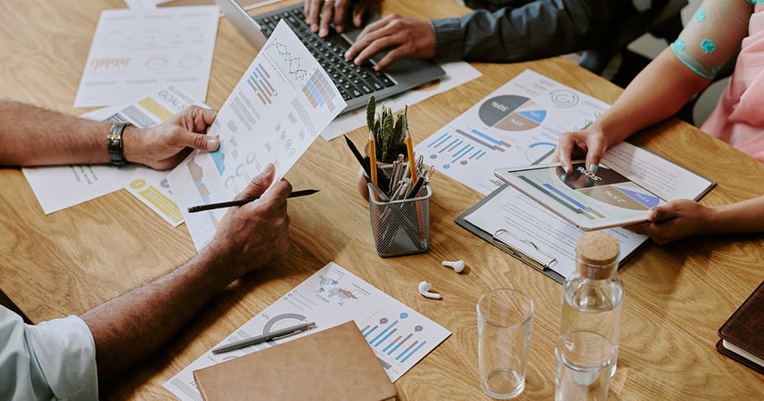 Hands holding a piece of paper filled with graphs and charts next to a desk filled with more printed pages filled with colorful graphs and charts