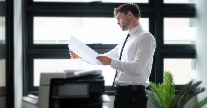man standing at an office printer looking at the color on a printed piece of paper