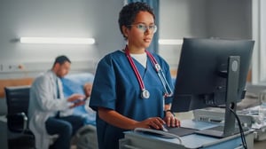 A health care worker entering information into a computer terminal. In the back ground, a provider is talking to a patient.