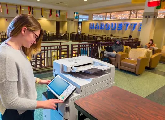 A student using a copy machine inside the Marquette Student Center. The work Marquette is in large letters in the background.