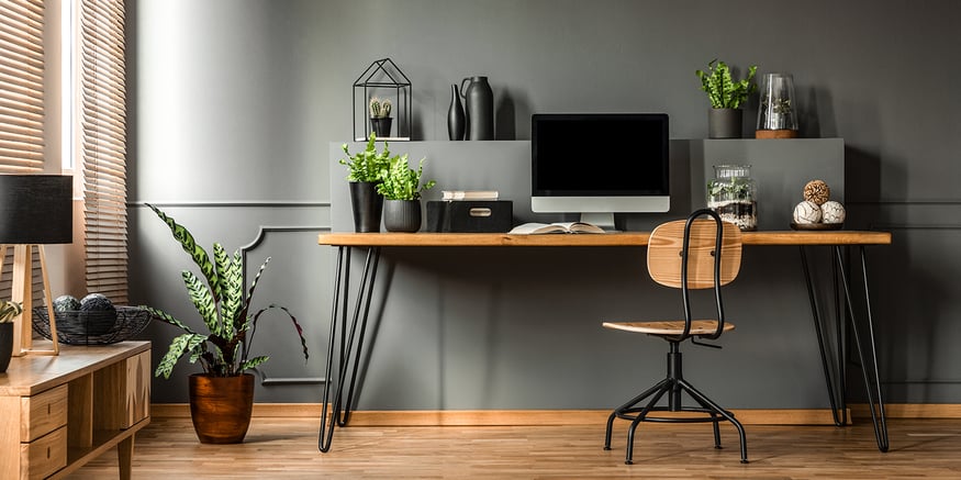 Desk with computer, plants, and other office items on a wooden floor with a wooden swivel chair.