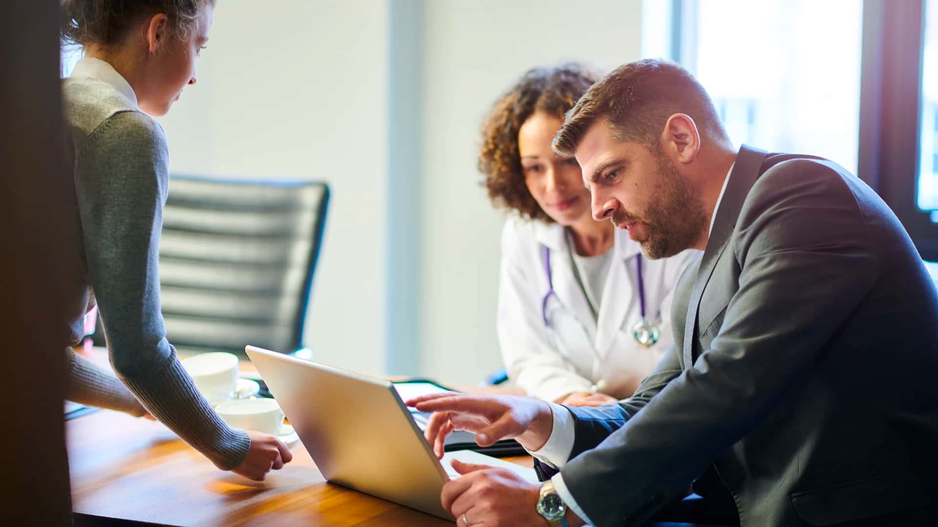 A person in a business suite reviewing IT files on a laptop with a person in a lab coat and stethoscope