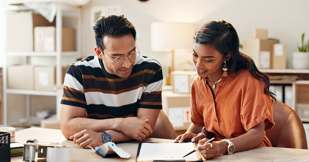 Two office workers sitting at a table discussing a written checklist attached to a clipboard