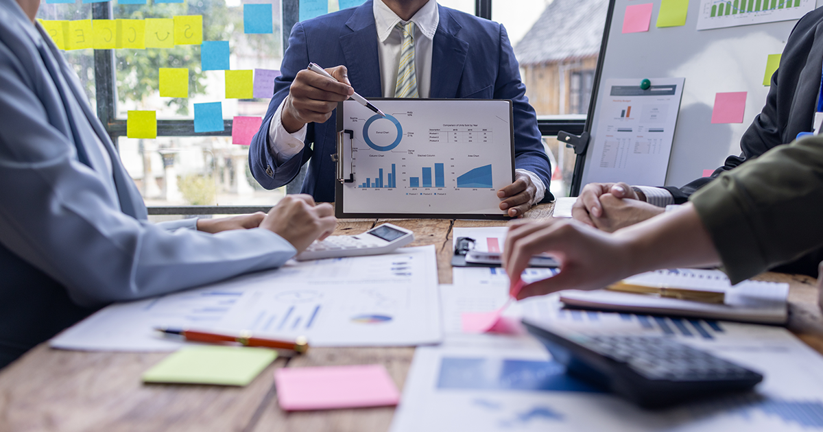 a man showing graphs and charts in a busy conference room table full of people and papers