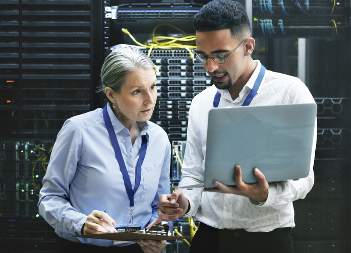 Two people standing in front of servers. looking at a laptop screen and talking